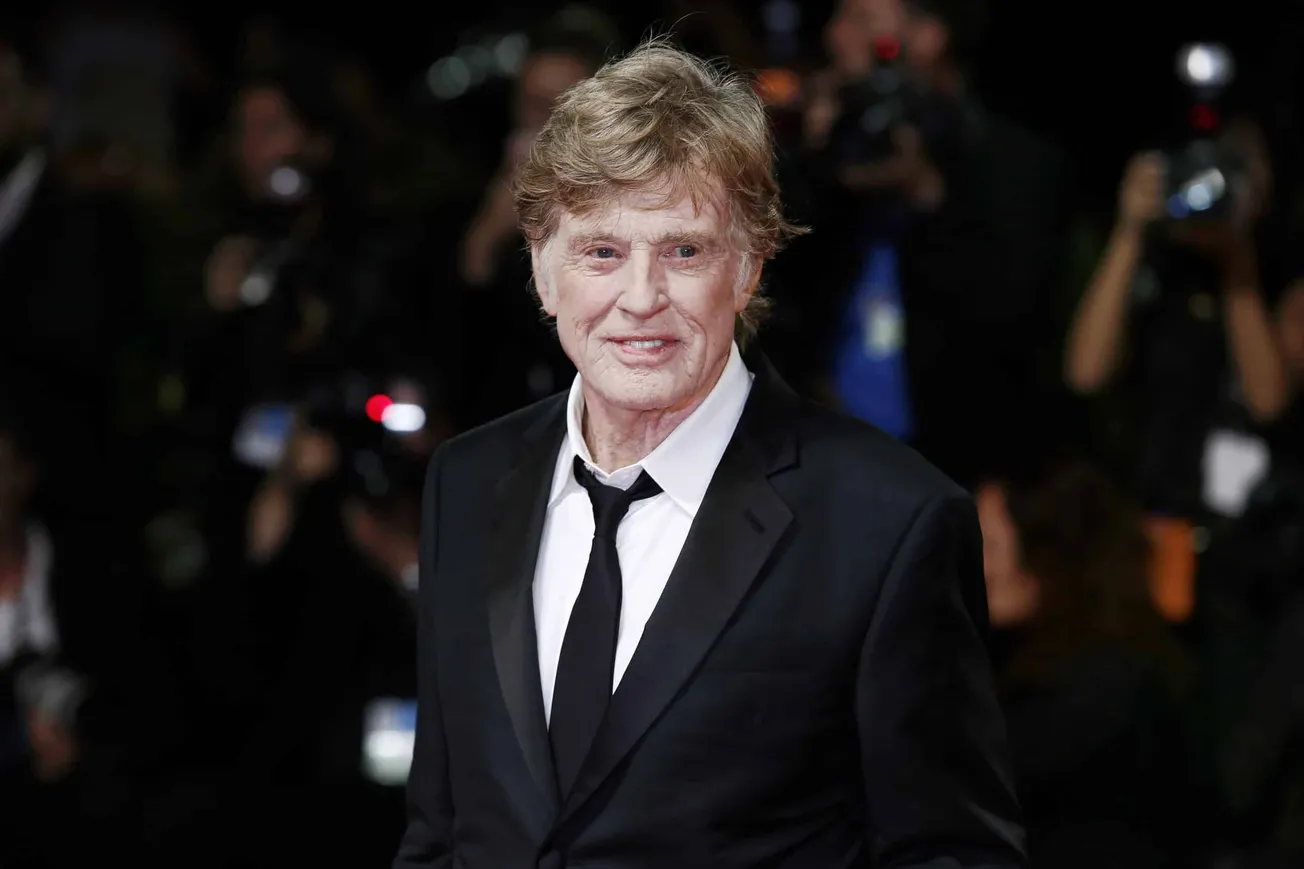 the late Robert Redford smiling at a red carpet event. He wears a dark suit and loose tie over a white shirt.