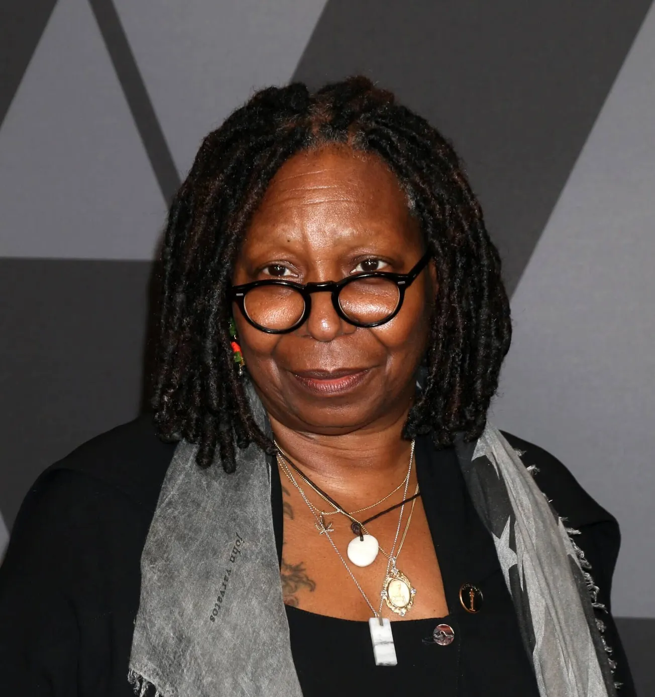 Whoopi Goldberg smiles at a press event, looking over her glasses.
