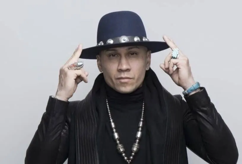 Taboo wears black and a black hat. His fingers are holding his hat. He wears a long piece of jewellery around his neck.