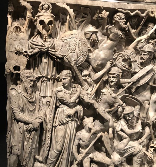 A carved stone sarcophagus in high relief showing a bearded man and woman crying in front of a trophy, next to a chaotic tangle of soldiers and horses in battle