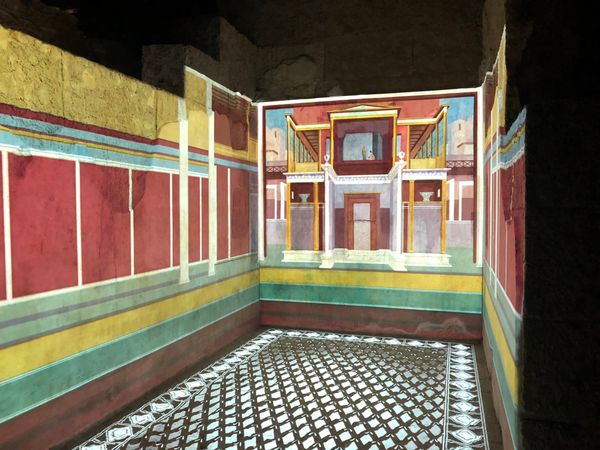 Lights projected on a dark concrete wall show red, green, and yellow wall painting and a black and white diamond pattern on the floor, with architectural designs on the far wall