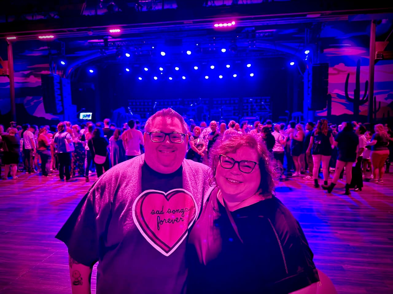 A man and woman take a photo in front of a stage in a music venue.