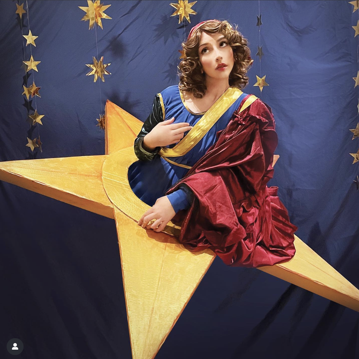 A woman cosplaying as the woman from the cover of the Mellon Collie and the Infinite Sadness album. She is wearing a blue dress with yellow trim, and a red cape on one shoulder. She is shown from the waist up, coming out of a large yellow star. She is on a blue background with smaller stars behind her.