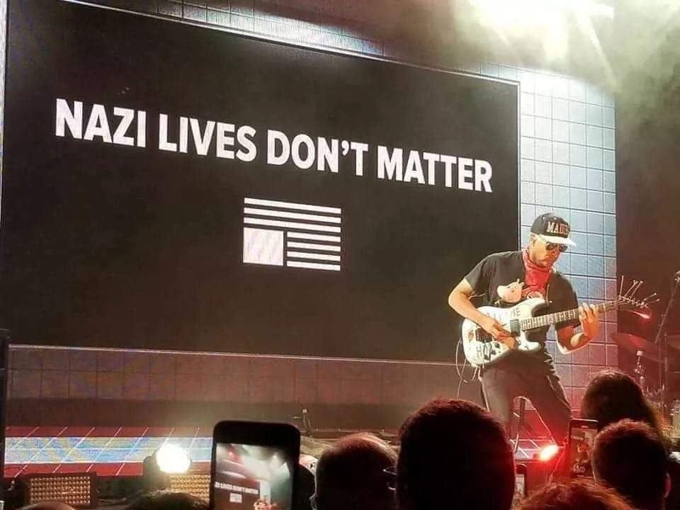 Tom Morello playing guitar on stage in front of a banner with an upside-down American flag with the text "NAZI LIVES DON'T MATTER."