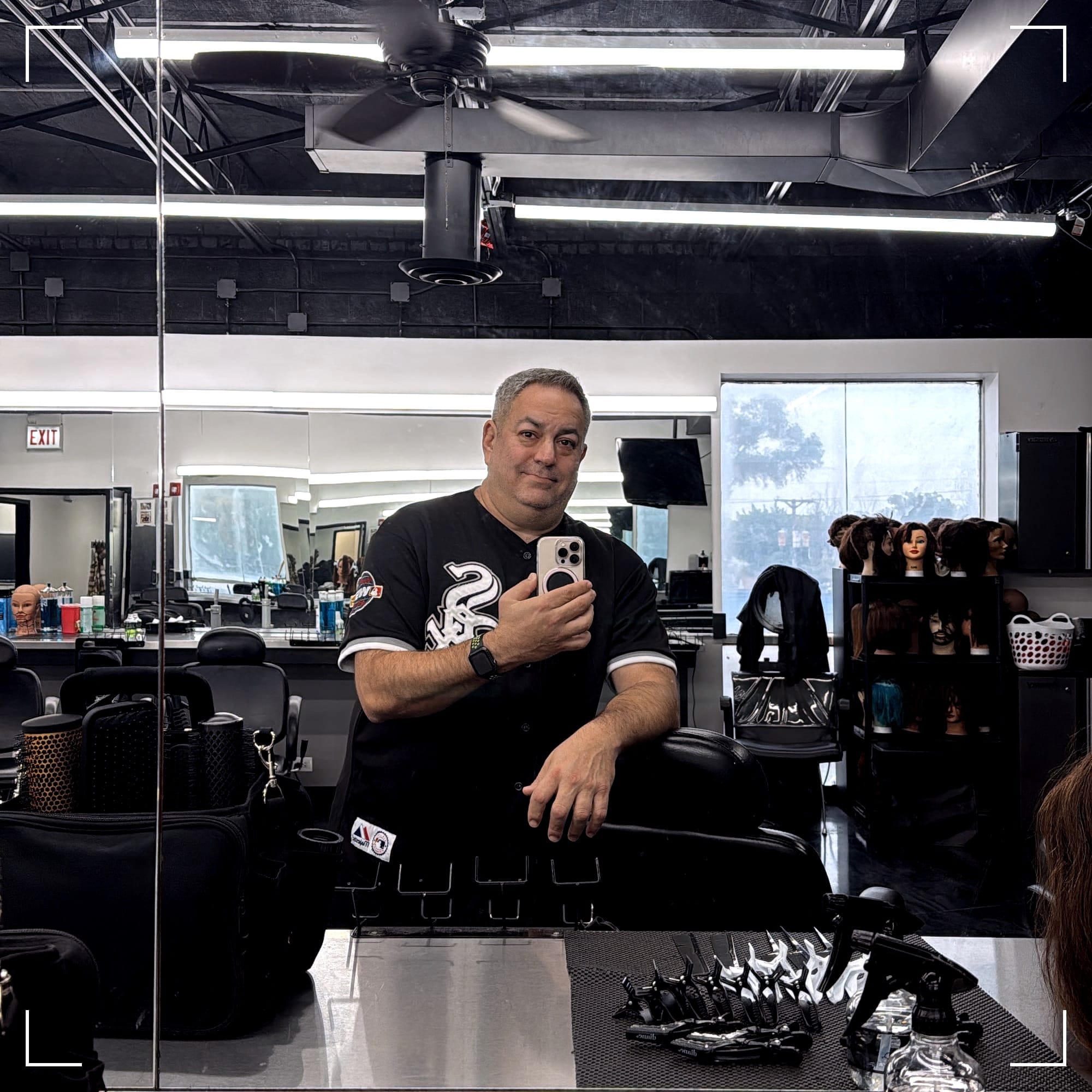 Aaron, holding his phone, taking a selfie in a mirror in front of his barber chair and station