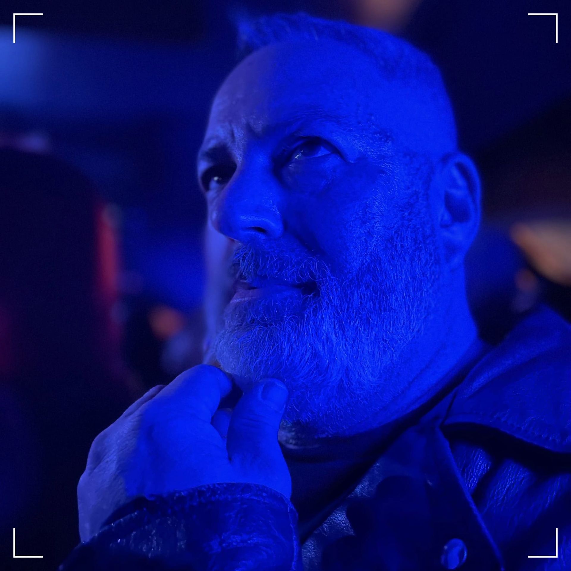 Aaron, wearing a biker jacket, hand on chin stroking his beard, in blue light