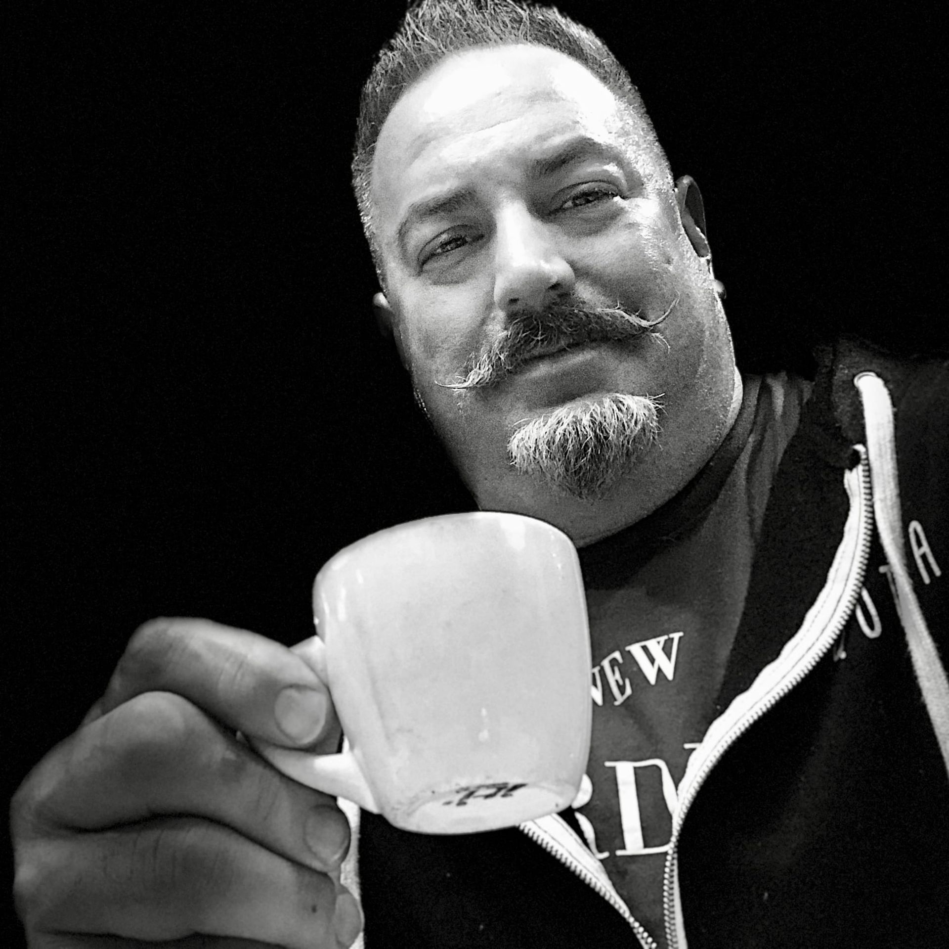 A black and white portrait of me, with a well groomed and waxed mustache and goatee, holding a cup of coffee.