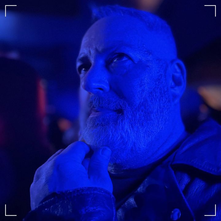 Aaron, wearing a biker jacket, hand on chin stroking his beard, in blue light