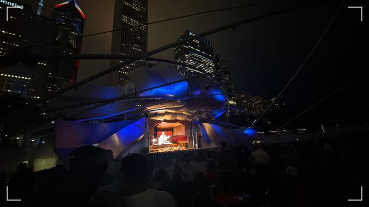 The Pritzker Bandshell/Pavilion in Chicago's Millennium Park, lit in blue at night against an overcast, hazy Chicago backdrop at night.