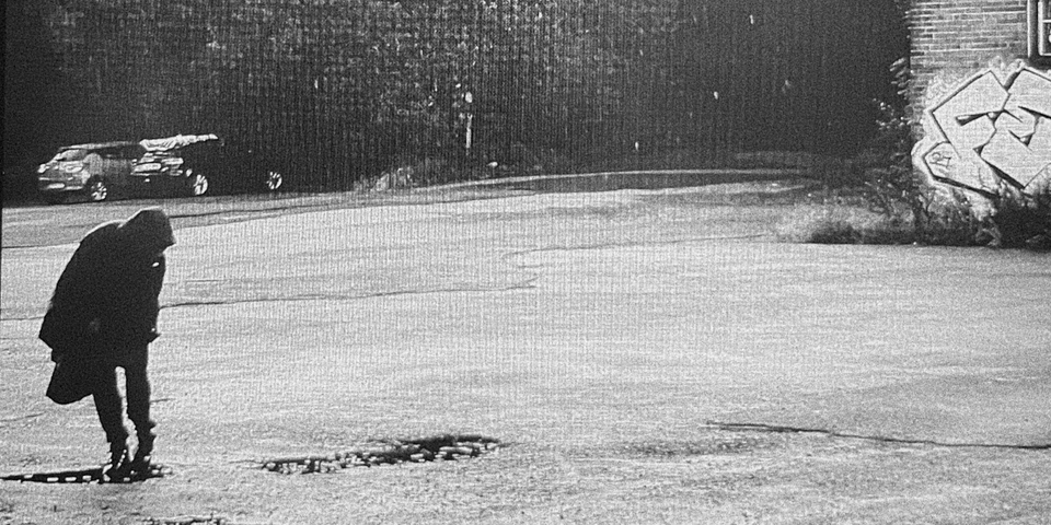 black and white crop of cover art, figure in long black coat on left of frame showing tarmac and trees, graffiti on a building edge of right frame