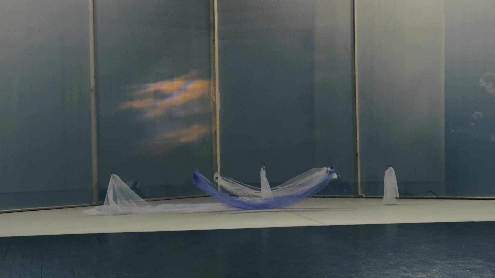 A stage with a red-blue cloud shimmering above a boat-like, semi-floating set of white and purple fabrics.