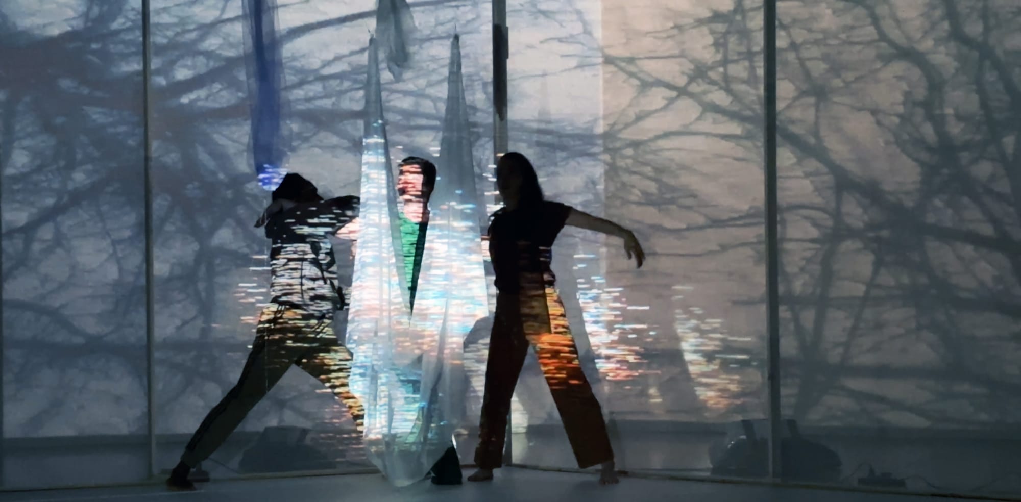 Three silhouettes of dancers before a projection of trees; the dancers are behind a fabric, all lit with a glowing digital glitch
