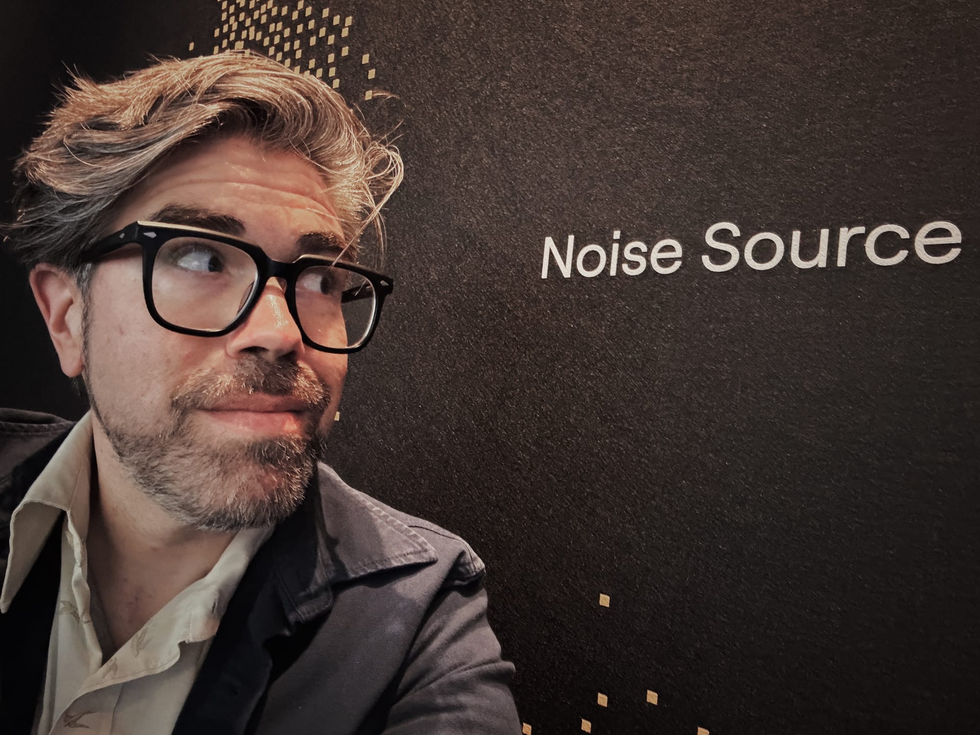 Man with beard and black glasses looking at “Noise Source” written on wall.