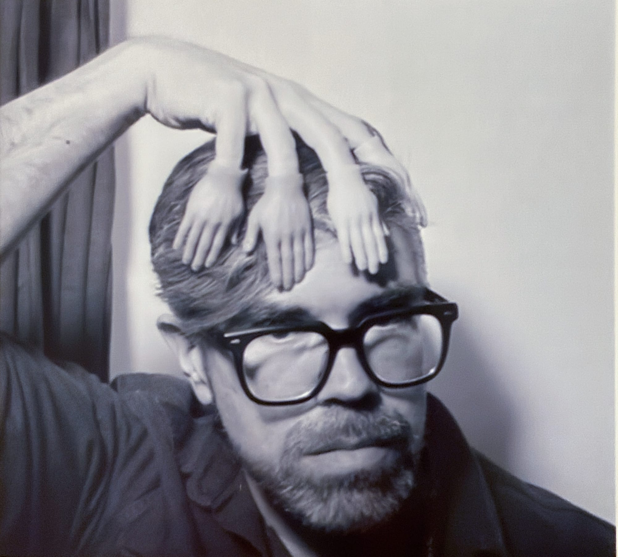 Film photograph of a bearded man in black glasses with his hand on his head, but his fingers have little plastic hands, as if he's from an AI generated image. 