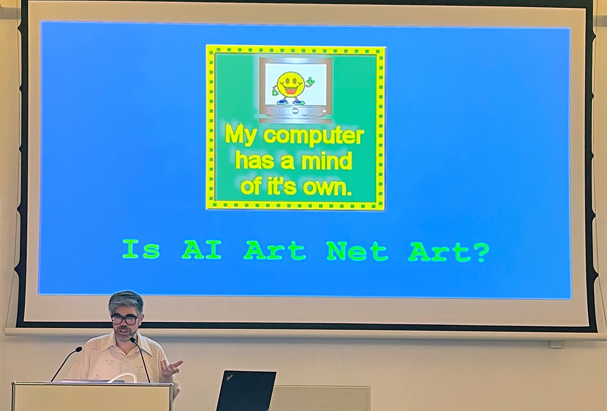 A bearded man with gray hair and glasses in front of a projection of a blue screen and a 90's era gif, a smiley face inside a computer with the text "My computer has a mind of it's own".