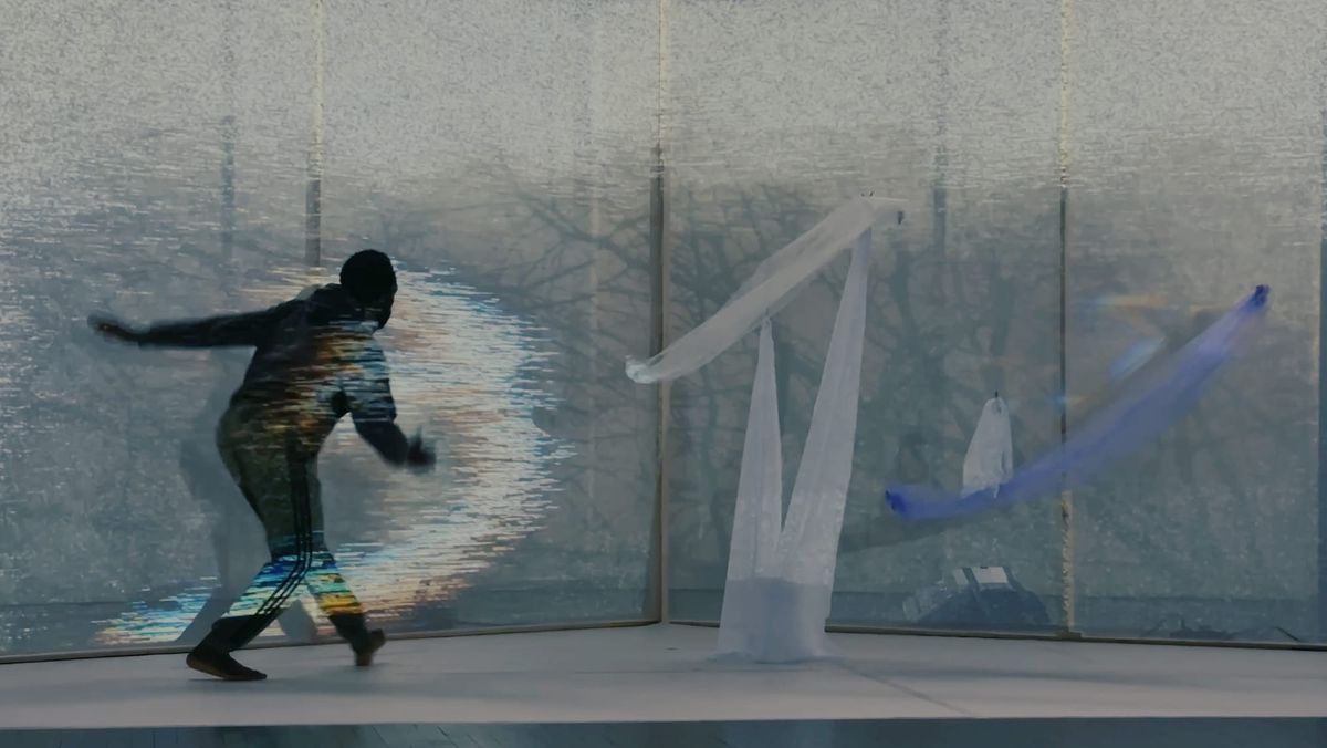 A dancer dressed in black emitting a streak of light against a backdrop of trees and floating fabrics.