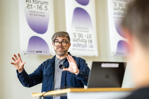 A bearded man with black glasses gestures with open hands at a podium in front of banners for the AI Forum, with purple blobs. 