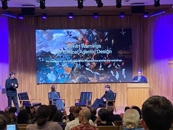 Two men standing on other side of a stage, Eryk Salvaggio at podium, two people seated. On screen: an image of a flock of birds captioned "Seven Warnings for Critical Agentic Design"