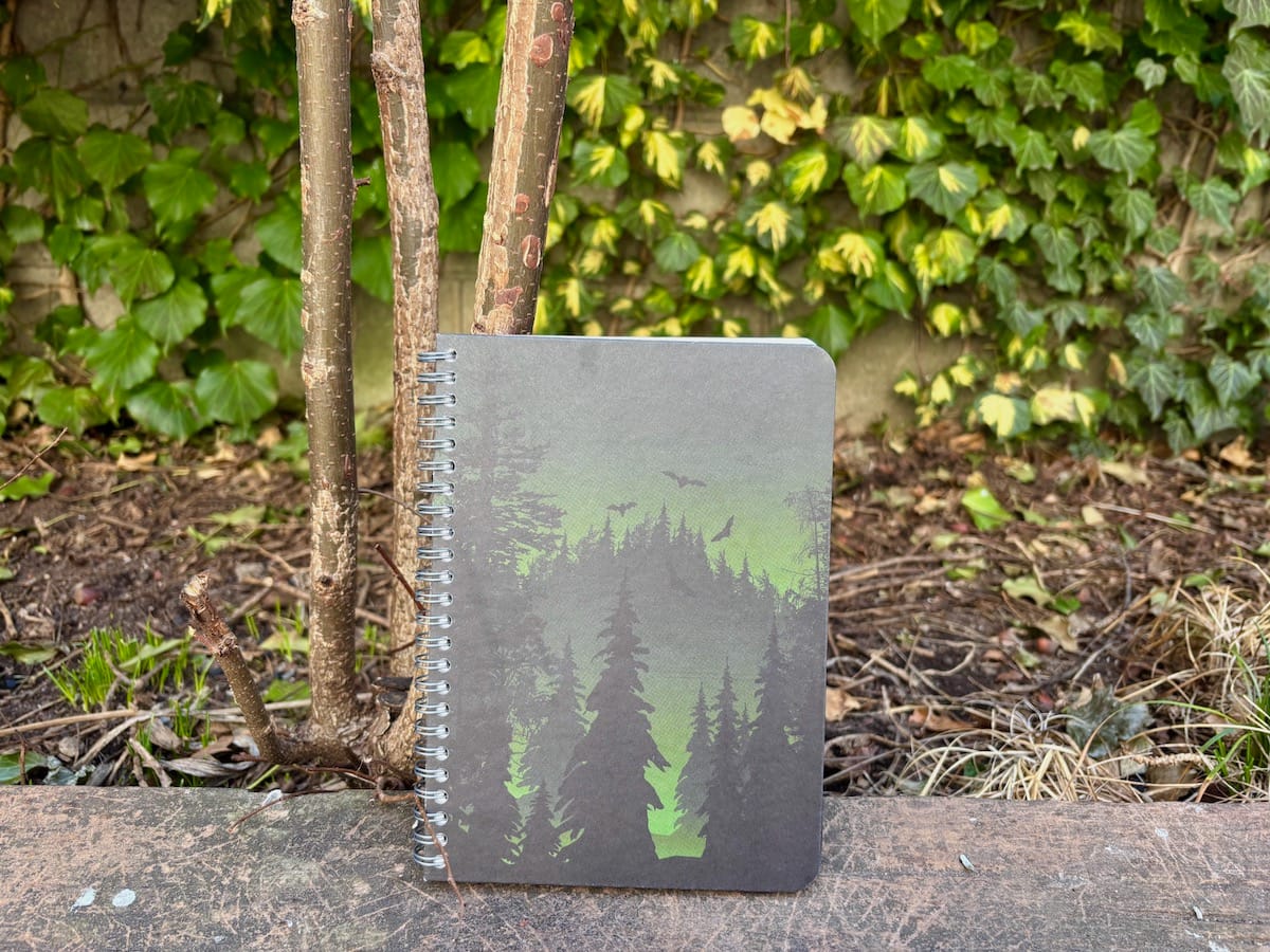 [A hardback green spiral notebook with a drawing of sillouetted dark pine trees over a green backdrop; bats—eek!—fly in the sky over the mountain. Behind the notebook is a peek at dirt and a wall of climbing green ivy.]