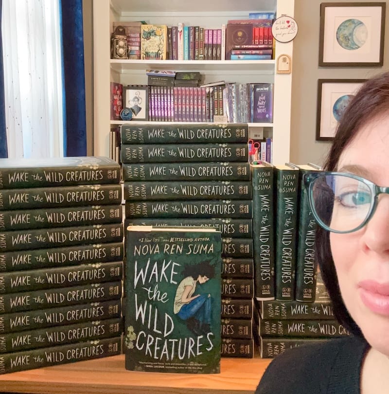 [A picture of stacks of green hardcovers of WAKE THE WILD CREATURES on a desk, with half the face of its author]