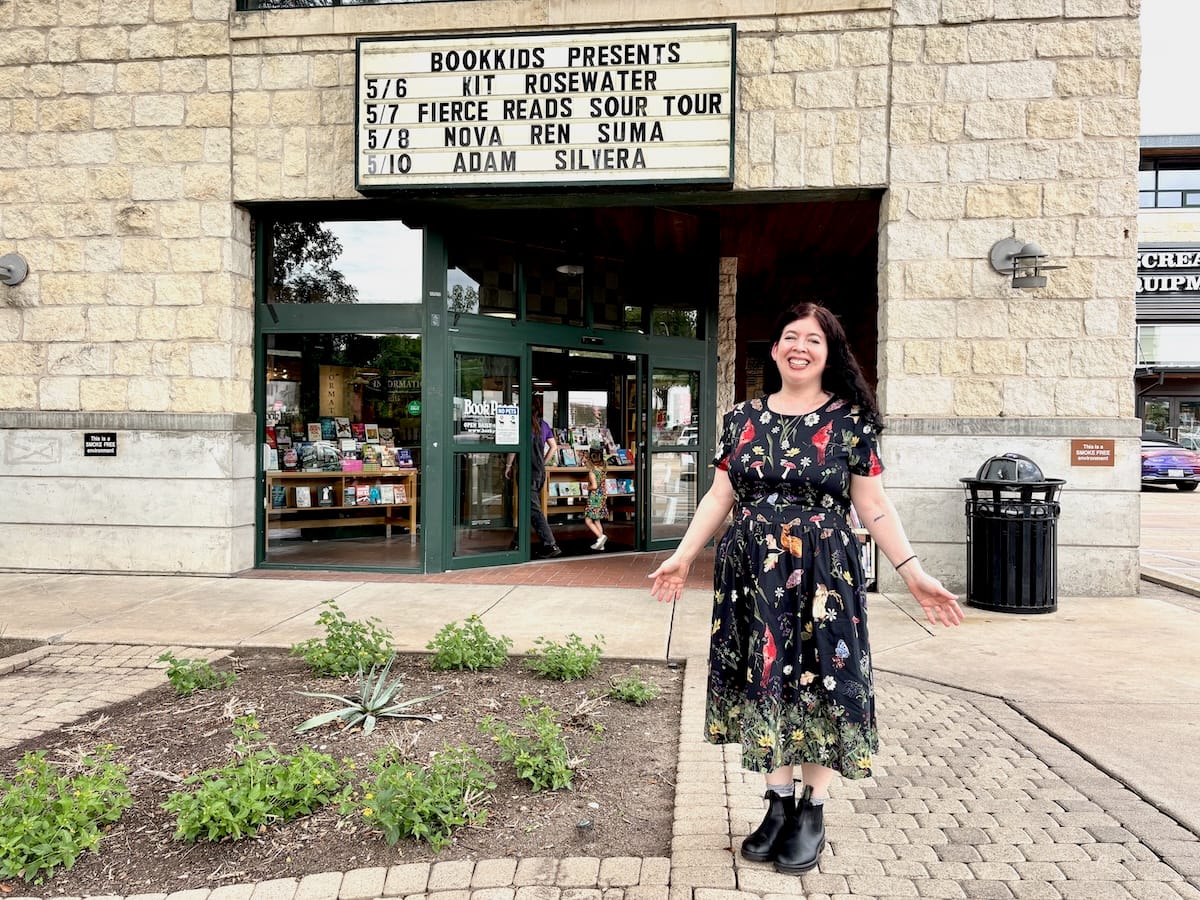 [A photo of me posing in a dress with plants and wild creatures all over it in front of the sign outside Book People in Austin, Texas. My name is on the sign!]