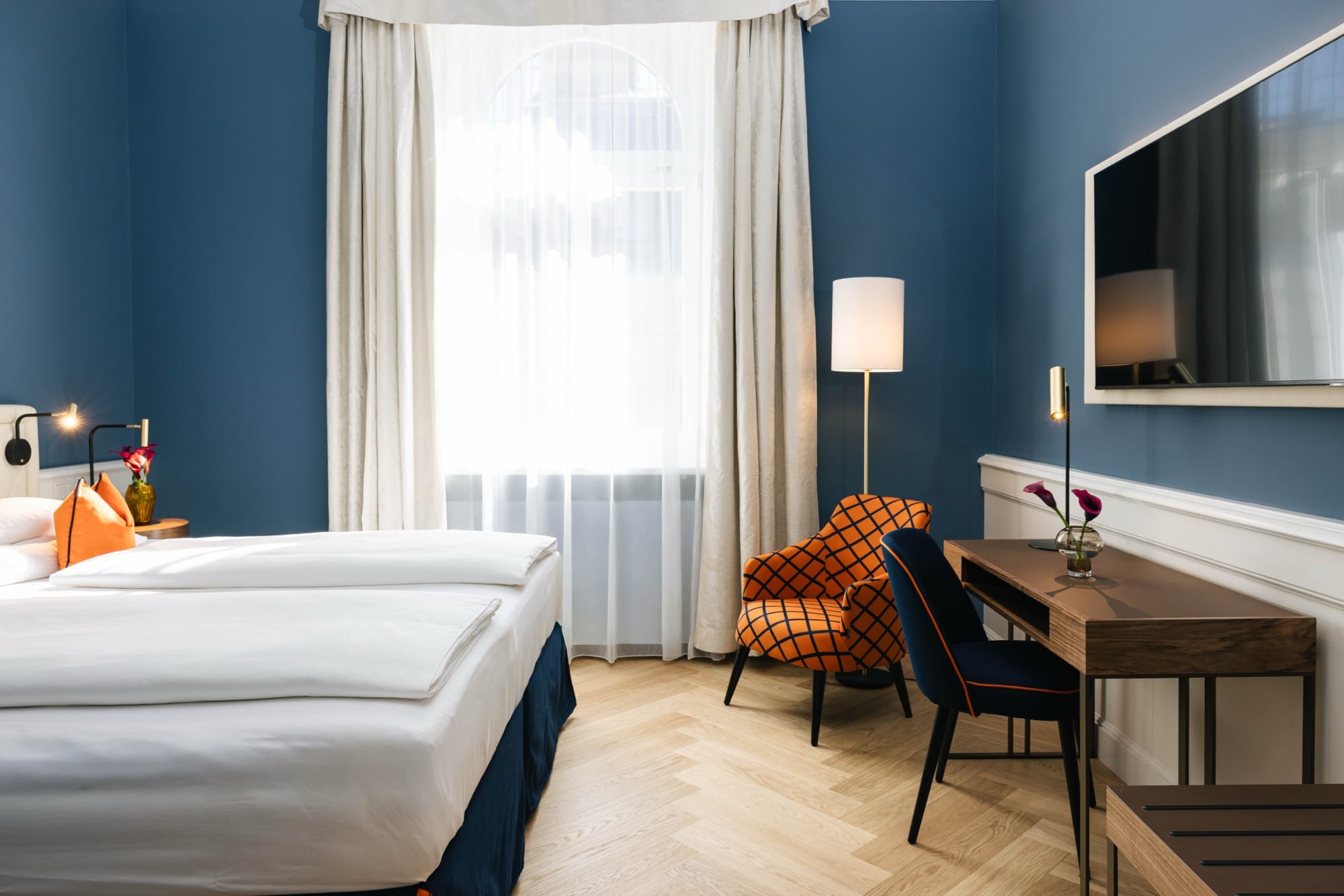 Hotelzimmer mit blauen Wänden, Doppelbett, Sessel mit Karomuster und Schreibtisch vor dem Fenster