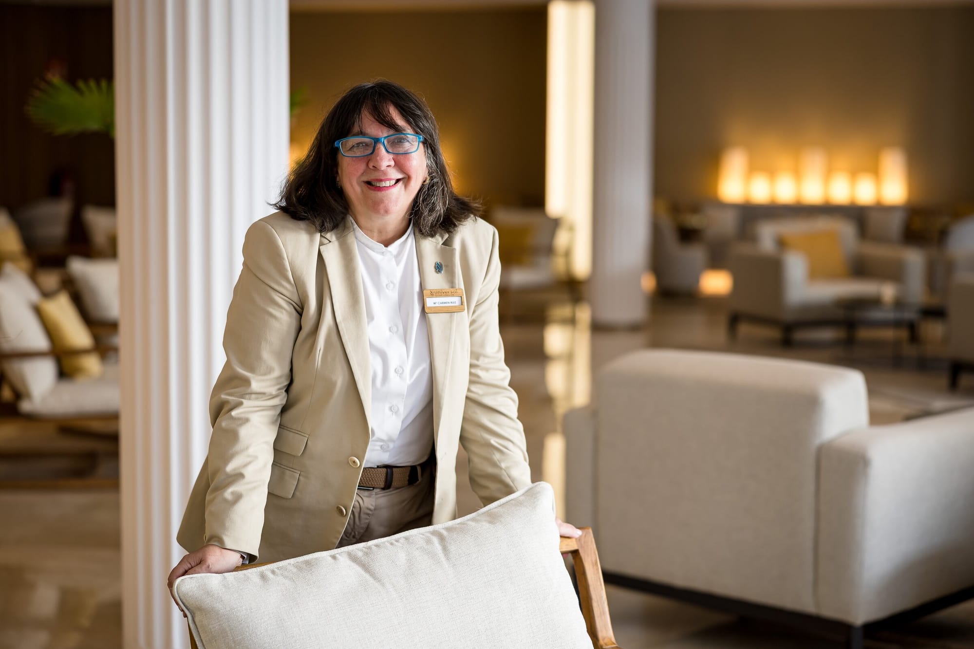 Maria del Carmen Ruiz Caro mit blauer Brille und beigem Blazer lehnt lächelnd an einem Sessel in der Hotellobby