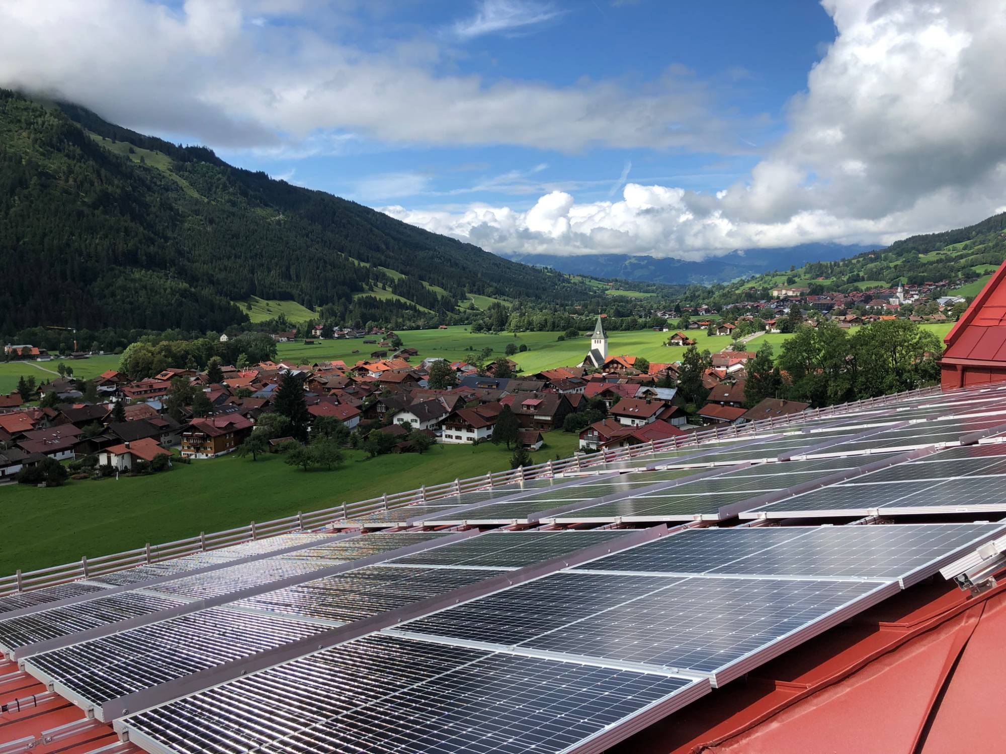 Photovoltaikmodule auf dem roten Dach des Hotels Prinz-Luitpold-Bad, dahinter ein Dorf mit Kirchturm im grünen Tal von Bad Hindelang