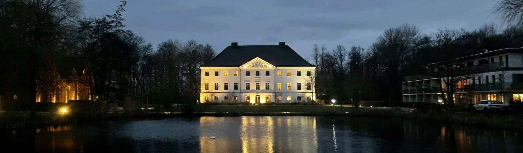 Das beleuchtete weiße Herrenhaus spiegelt sich in der Dämmerung im Teich