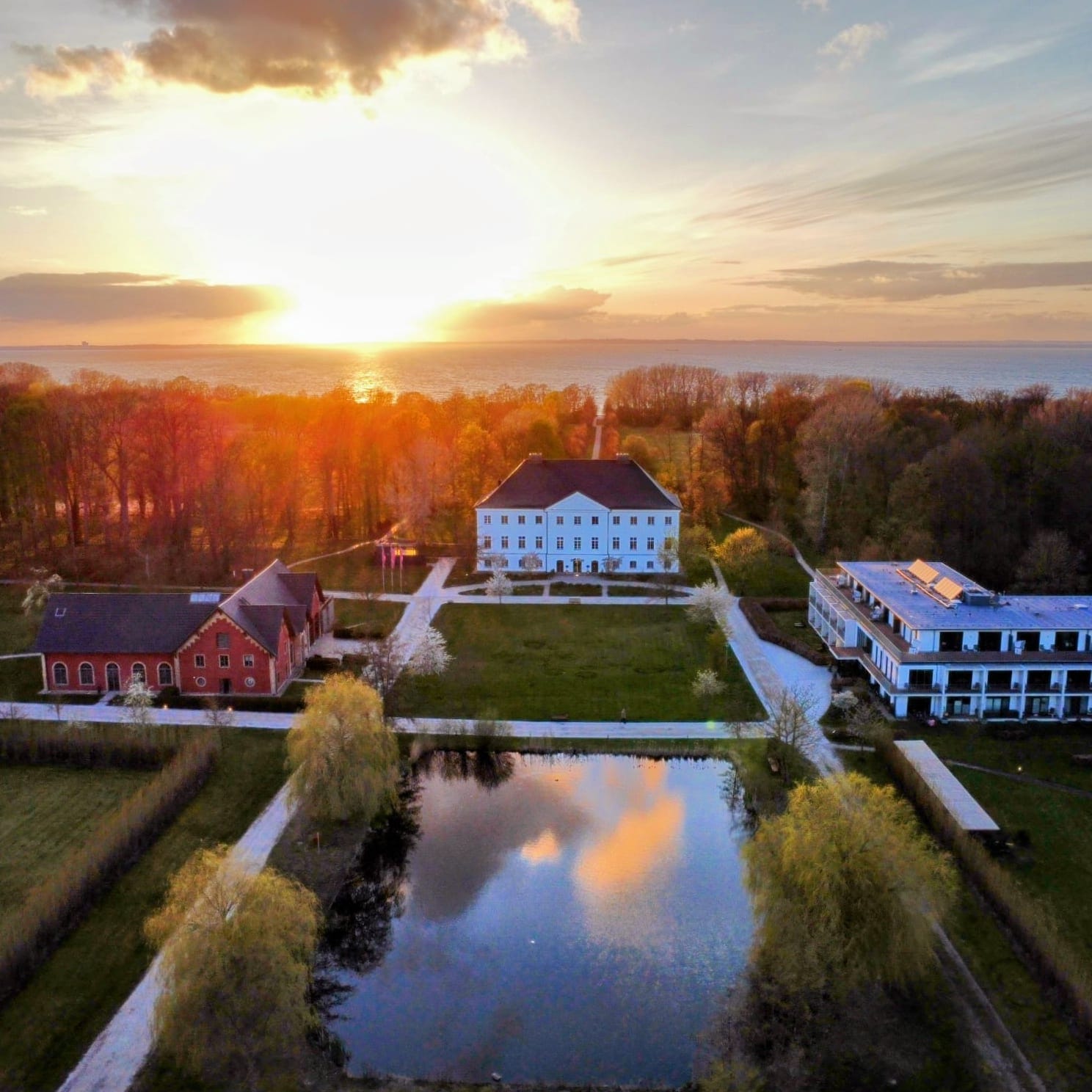 Luftaufnahme des Schlossguts mit weißem Herrenhaus, roter Backsteinremise, modernem Hotelbau und rechteckigem Teich, dahinter die Ostsee im Sonnenuntergang