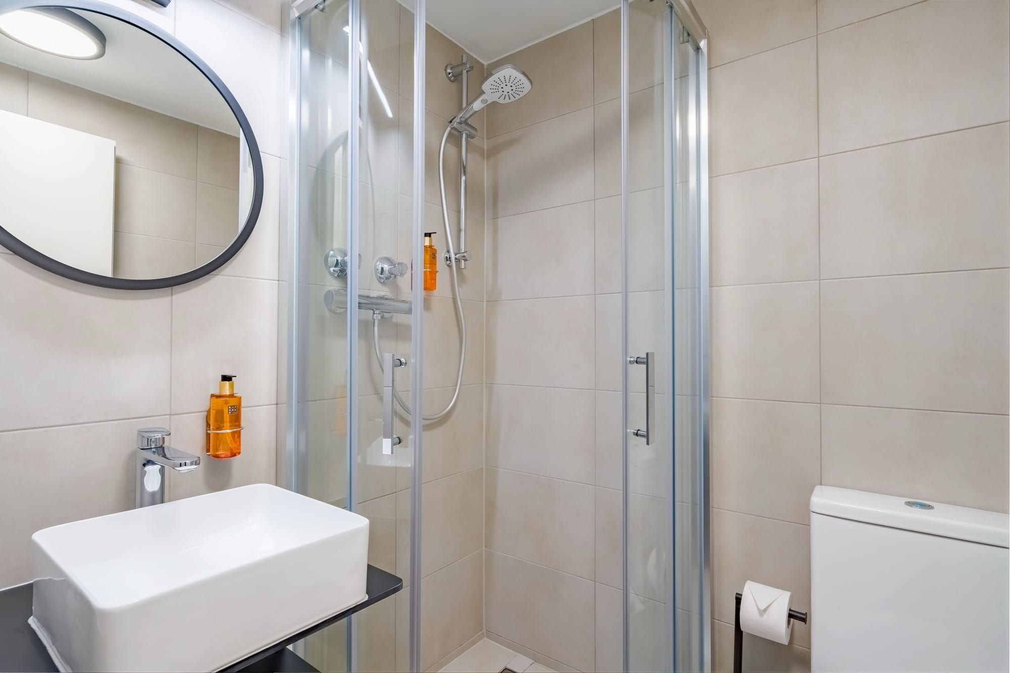 Hotelbad mit gläserner Eckdusche, rechteckigem Aufsatzwaschbecken, rundem Spiegel und WC