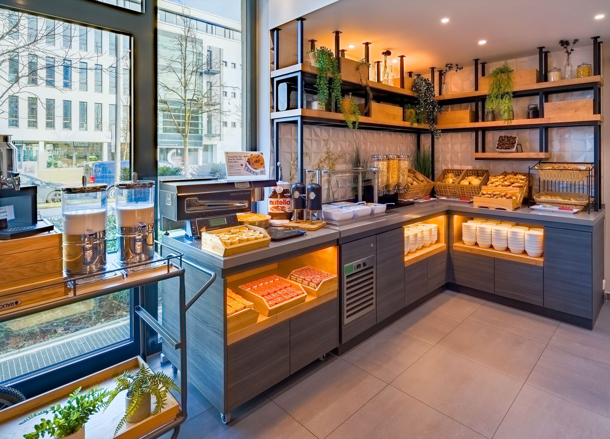 Frühstücksbuffet mit Brötchenkörben, Müslispendern, Saftbehältern und Holzregalen vor einer Fensterfront