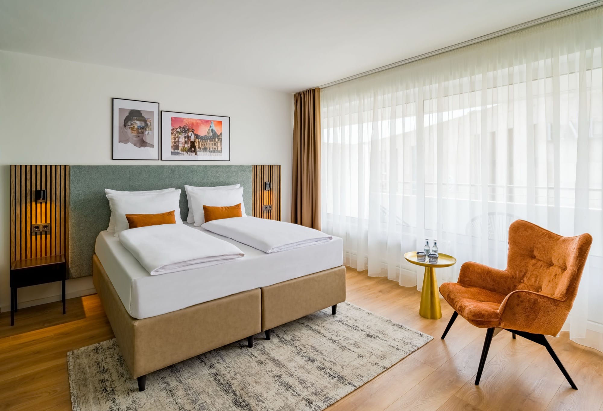 Hotelzimmer mit Doppelbett, orangefarbenem Ohrensessel, goldenem Beistelltisch und bodentiefen Fenstern mit weißen Gardinen