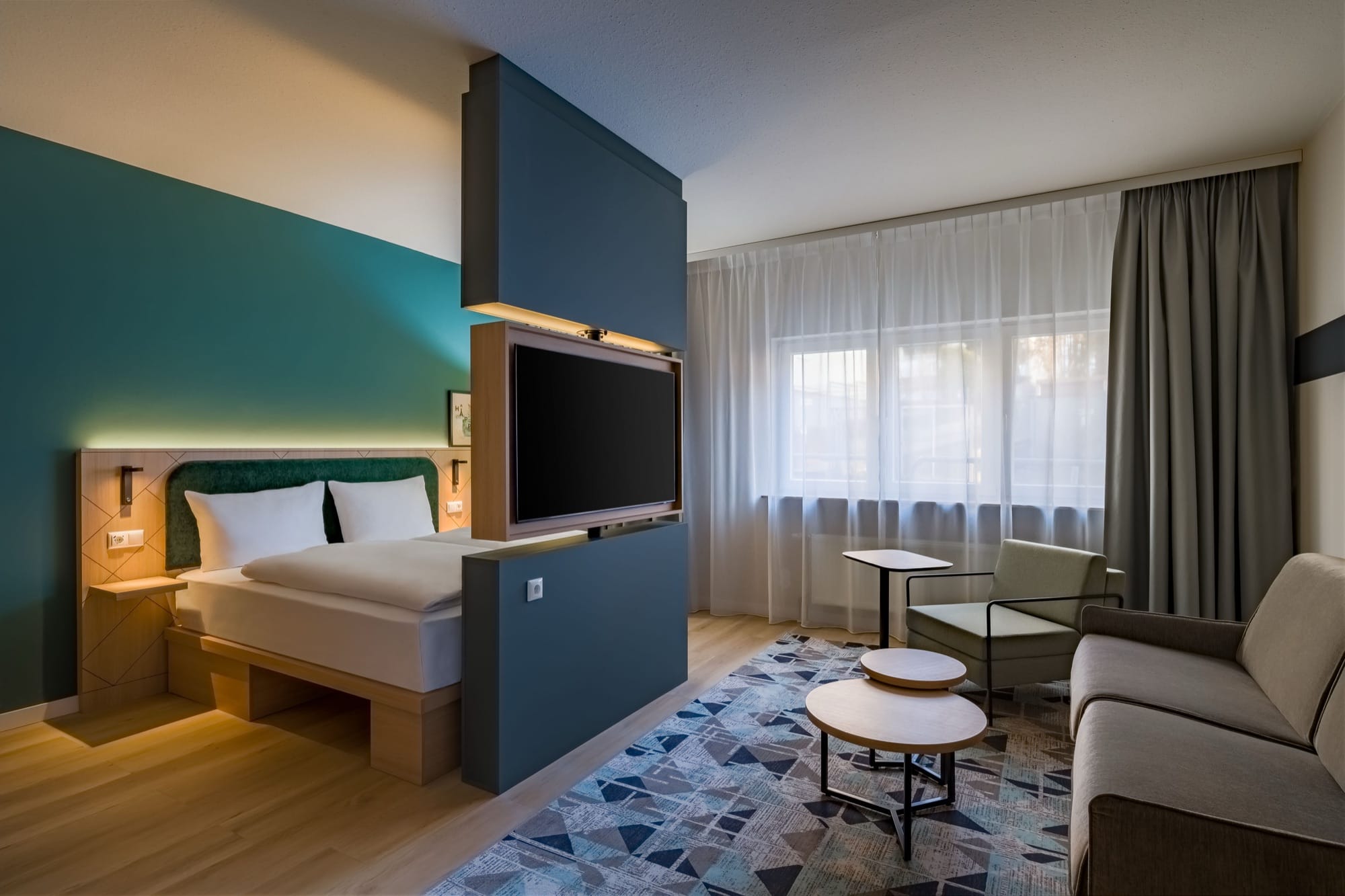 Hotelzimmer mit petrolfarbener Wand, Doppelbett, frei stehender Wand mit Fernseher, Sessel, Sofa und runden Couchtischen vor einem Fenster mit Vorhängen