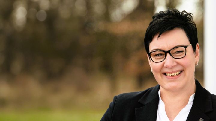 Janet Schroeder smiling, wearing glasses, a black blazer and a white blouse in front of a blurred park
