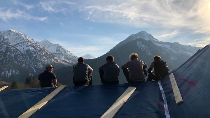 Five tradesmen sit on the ridge of the newly covered hotel roof, looking out at snow-capped Allgäu mountains