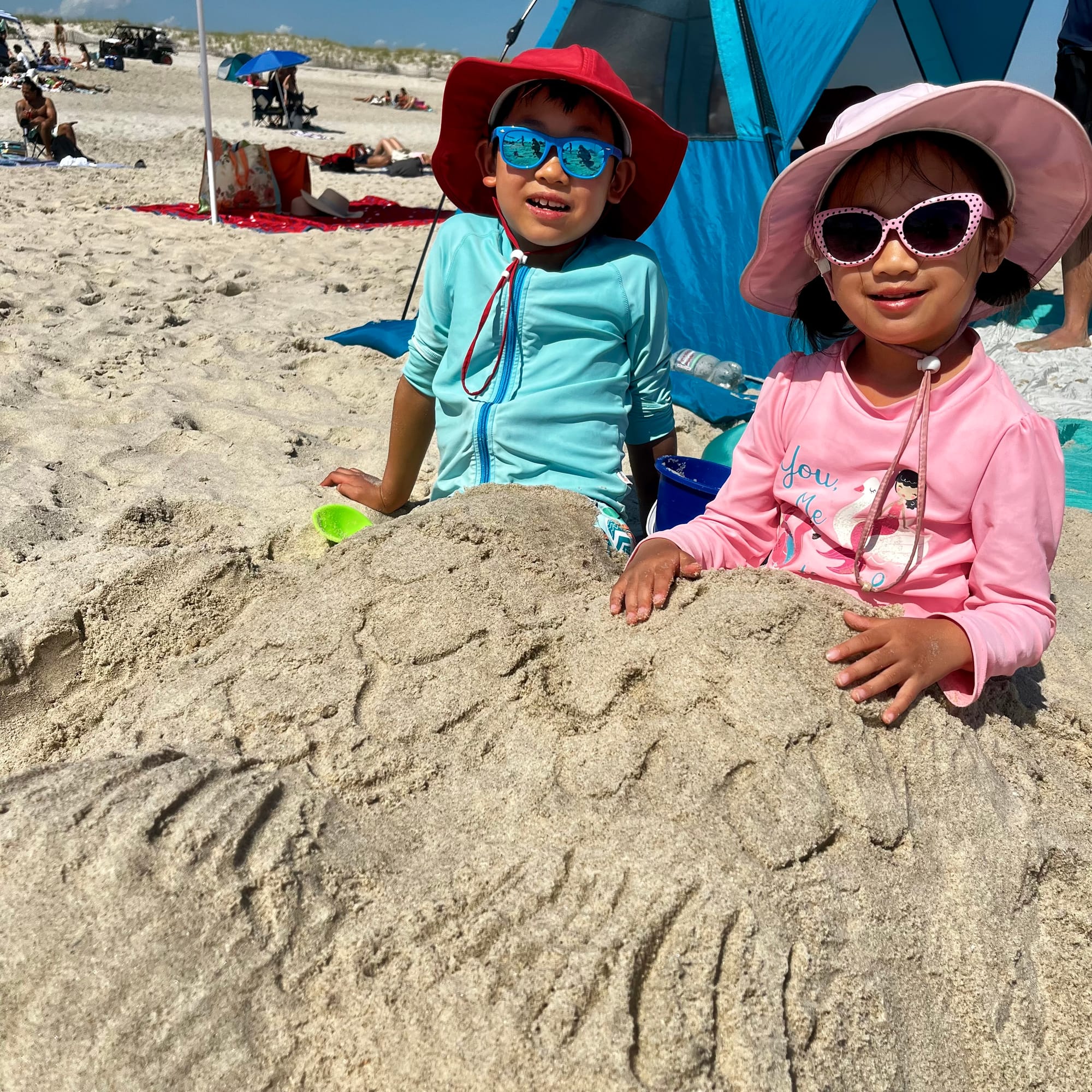 Two kids, boy in red hat, blue rashguard and blue sunglasses, girl in pink hat, shirt and sunglasses. Buried from waist down in sand with a mermaid tale design