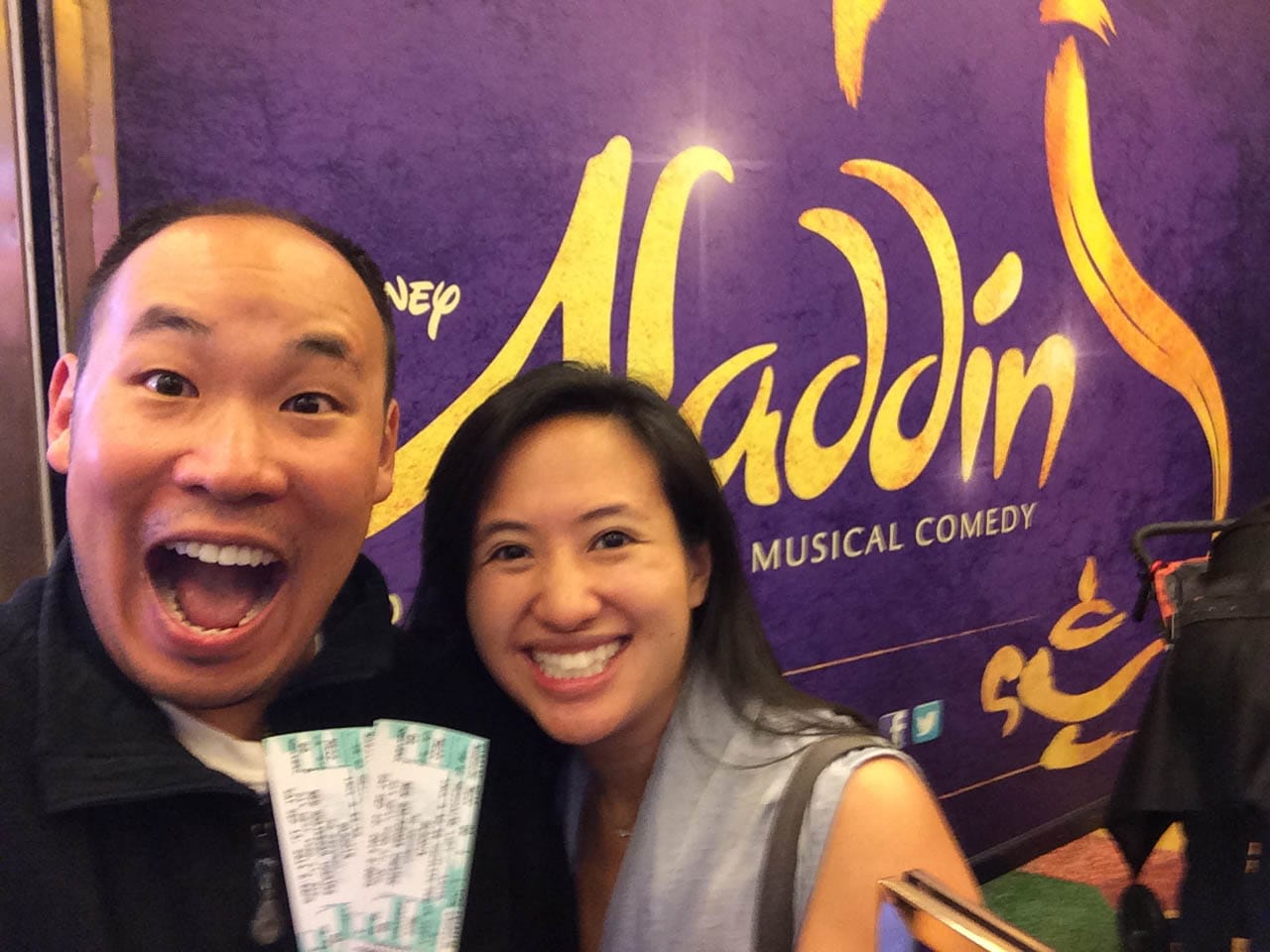 Husband and I in front of Aladdin Musical poster with two tickets and huge smiles