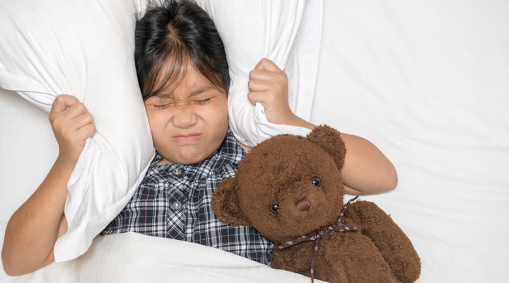 Little girl in bed covering ears with pillow to block out too loud noise and holding teddy bear 