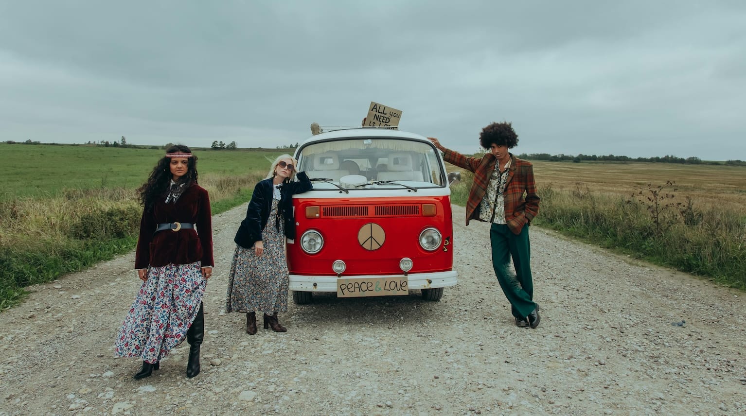 Three young people dressed in retro 1970s hippy style stand beside a red camper van with a peace sign on the front.