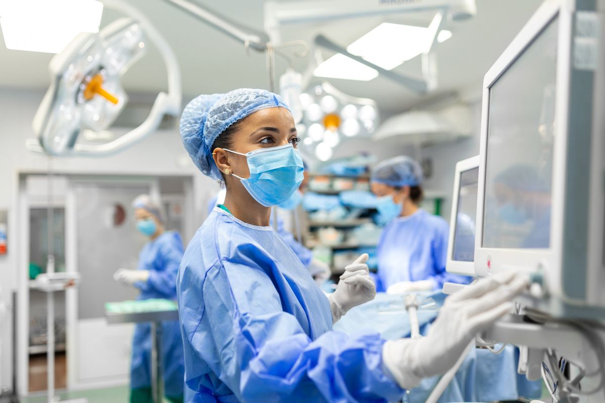 Anesthetist Working In Operating Theatre Wearing Protecive Gear checking monitors while sedating patient before surgical proc