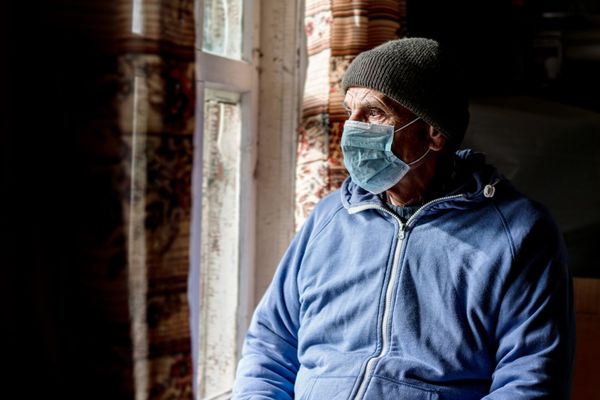 Aged male person wearing facemask and hat looking through window glass indoor
