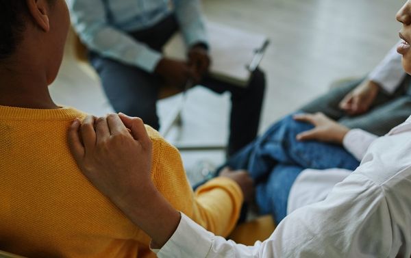 Close-up of hand of schoolgirl on shoulder of worried classmate sitting among his friends and psychologist and describing his