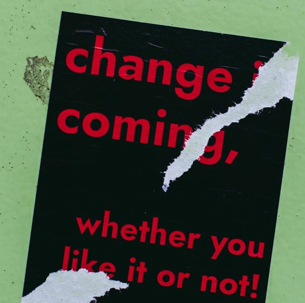 Green background with black sign, with red letters reading "Change is coming whether you like it or not"