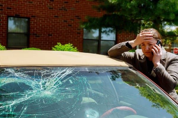 Woman on phone next to car with cracked windshield after street parking damage.