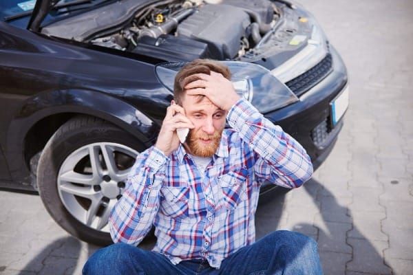 Man sitting on the ground beside a disabled car with the hood open, talking on his phone and looking stressed after a vehicle breakdown or accident.