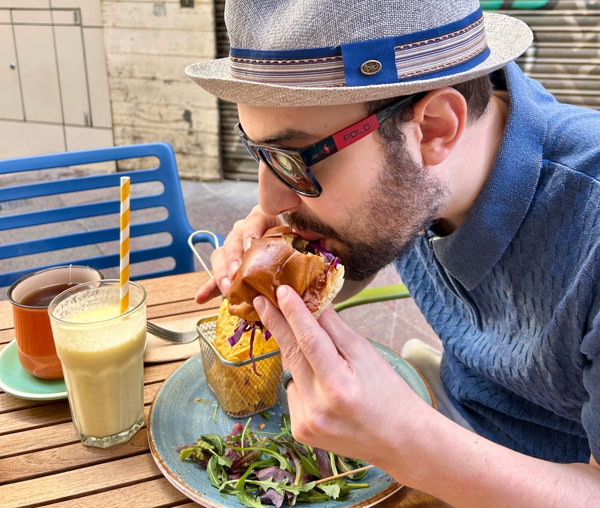 Juan in an alley, biting into a huge burger with a smoothie, cup of tea, and salad also in front of him.