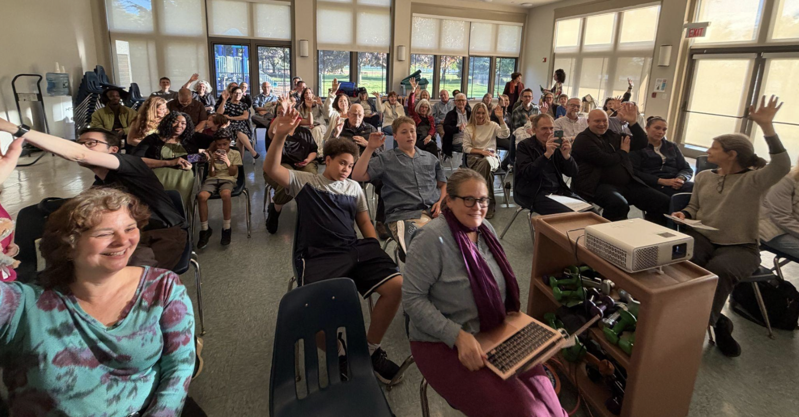 A crowd of all ages raising their hands and smiling at the Rivertowns Earth Month scavenger hunt awards event in a community room.