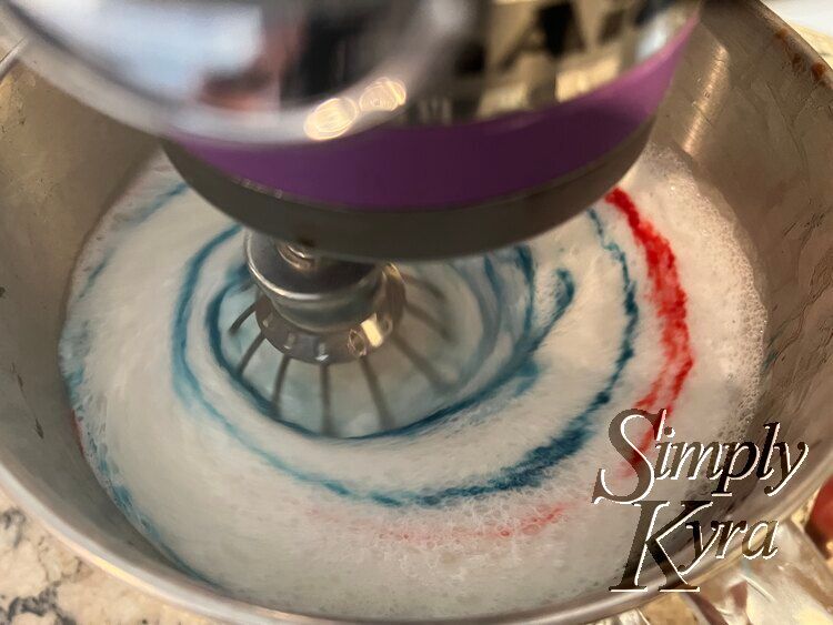Image shows the bowl of a stand mixer looking down with the whisk down, turned on, and blurred. The blue is streaked around several times while the red only looks streaked around once. 