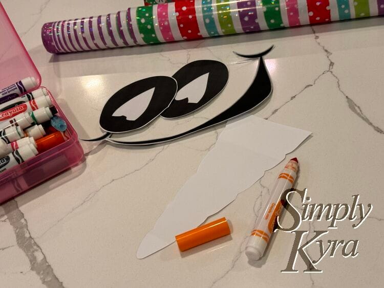 Image shows the cut out eyes and mouth, the wrapping paper, markers, and cut the paper carrot.